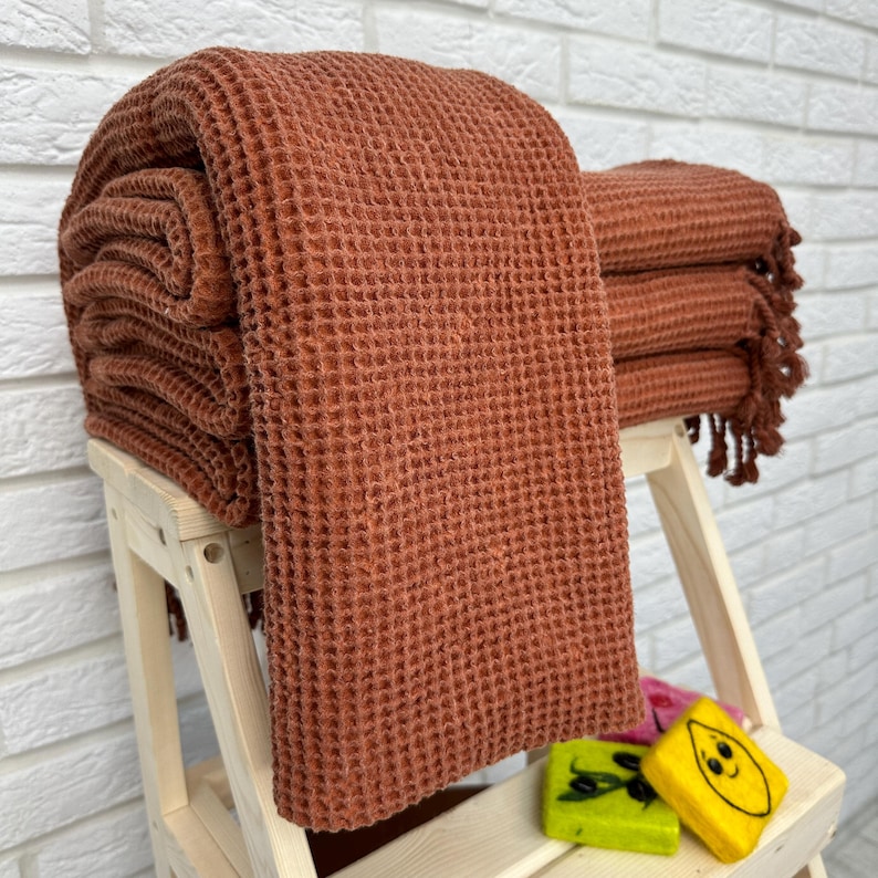May include: A stack of terracotta waffle weave towels with fringed edges. The towels are folded and displayed on a wooden step stool.