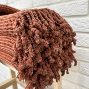 May include: A rust-colored woven throw blanket with a fringe border. The blanket is folded and stacked on a wooden surface.