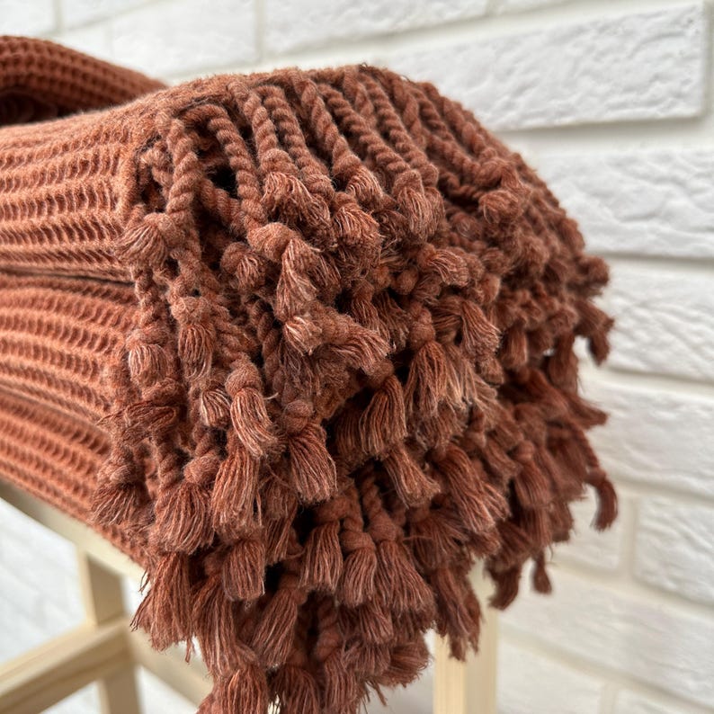 May include: A rust-colored woven throw blanket with a fringe border. The blanket is folded and stacked on a wooden surface.