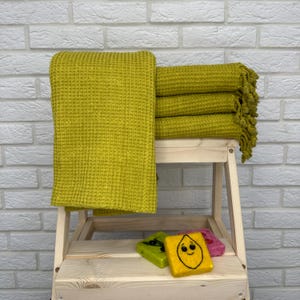 Peut inclure: Une pile de quatre serviettes en nid d'abeille jaunes avec des bords frangés. Les serviettes sont pliées et empilées sur un tabouret en bois. Il y a trois petites décorations en feutre sur le tabouret, dont un citron jaune avec un visage souriant.