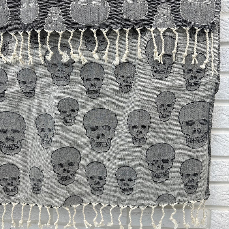 May include: A gray and white patterned fabric with a fringe border. The fabric features a repeating pattern of black skull designs.