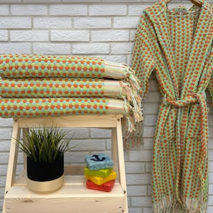 May include: A stack of four green and orange towels with a fringe border. The towels are folded and stacked on a wooden shelf. A green and orange hooded robe with a fringe border is hanging on a white brick wall.