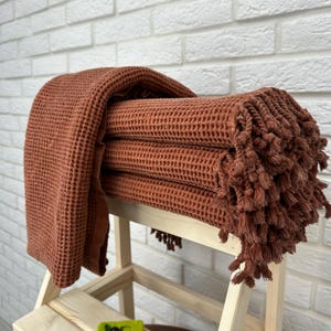 May include: A stack of four rust-colored waffle weave towels with fringe trim. The towels are folded and displayed on a wooden stool.