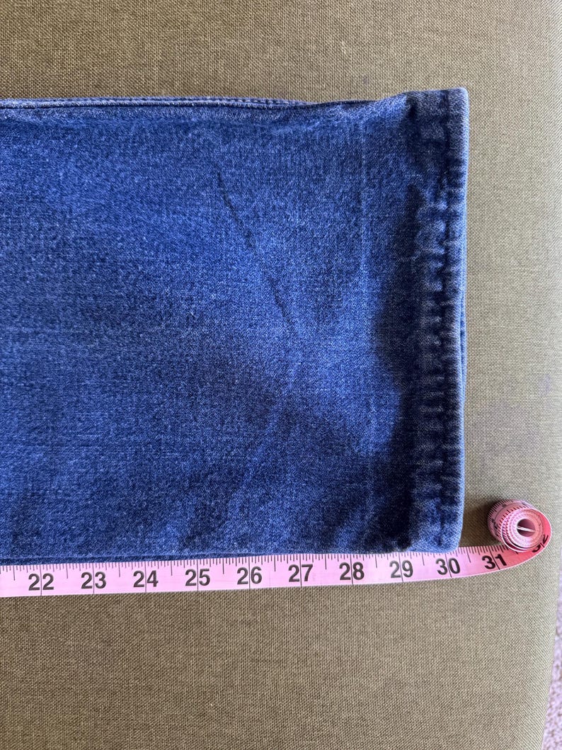 May include: Close-up of blue denim fabric with a pink measuring tape showing a length of 30 inches. The denim has a textured appearance and is likely part of a pair of jeans.