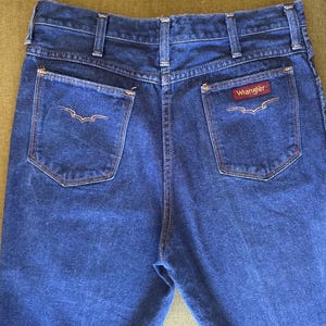 May include: Dark blue denim Wrangler jeans. The jeans have two back pockets with decorative stitching and the Wrangler logo on the right pocket. The jeans have a classic five-pocket design and a straight leg cut.