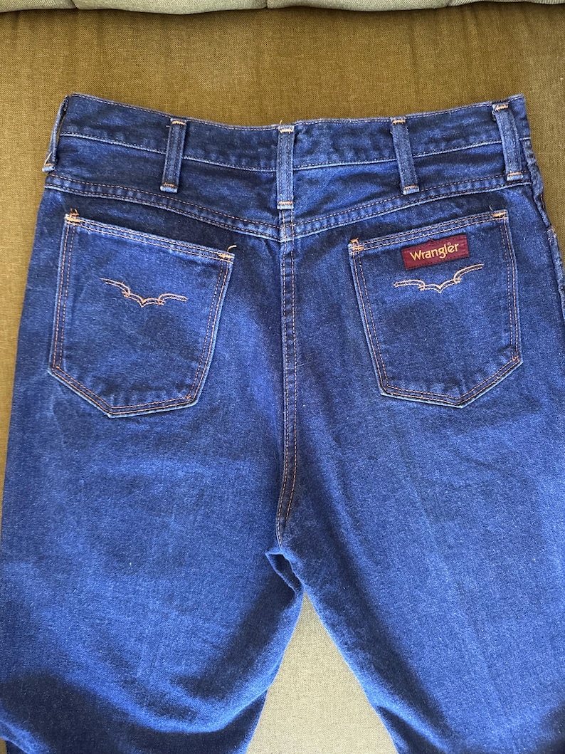 May include: Dark blue denim Wrangler jeans. The jeans have two back pockets with decorative stitching and the Wrangler logo on the right pocket. The jeans have a classic five-pocket design and a straight leg cut.