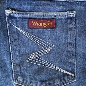May include: Close-up of a blue denim jean pocket. The pocket features white stitching in a zig-zag pattern and a rectangular maroon label with the brand name "Wrangler" in gold lettering.