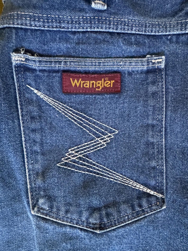 May include: Close-up of a blue denim jean pocket. The pocket features white stitching in a zig-zag pattern and a rectangular maroon label with the brand name "Wrangler" in gold lettering.