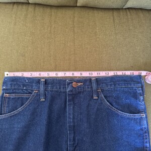 May include: Close-up of a pair of dark blue denim jeans. The jeans have a button closure, front pockets, and belt loops. A pink measuring tape shows the waist is approximately 16 inches wide.