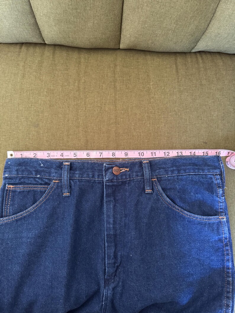 May include: Close-up of a pair of dark blue denim jeans. The jeans have a button closure, front pockets, and belt loops. A pink measuring tape shows the waist is approximately 16 inches wide.