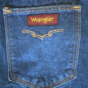 May include: Close-up of a dark blue denim jean pocket with orange stitching. A rectangular maroon patch with the word "Wrangler" in gold lettering is centered on the pocket. Below the patch is a decorative stitched design.