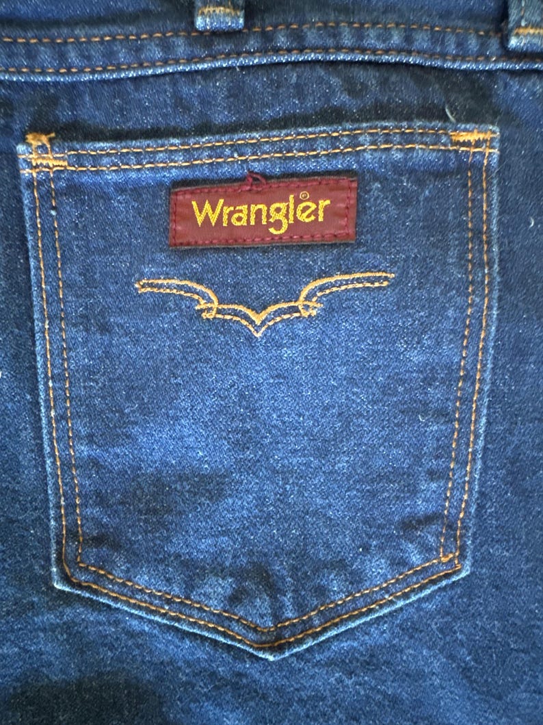 May include: Close-up of a dark blue denim jean pocket with orange stitching. A rectangular maroon patch with the word "Wrangler" in gold lettering is centered on the pocket. Below the patch is a decorative stitched design.