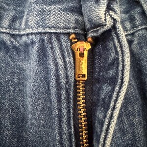 May include: Close-up of a blue denim garment with a brass-colored zipper. The zipper is partially closed, revealing the Talon brand name. The denim fabric shows visible texture and stitching details.