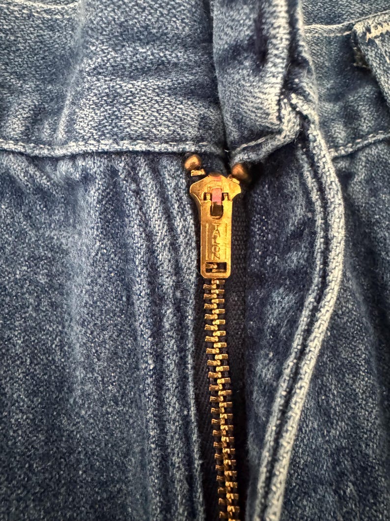 May include: Close-up of a blue denim garment with a brass-colored zipper. The zipper is partially closed, revealing the Talon brand name. The denim fabric shows visible texture and stitching details.