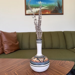 Może przedstawiać: Biały ceramiczny wazon z wąską szyjką i pękatą podstawą, zdobiony geometrycznym wzorem z południowego zachodu w odcieniach pomarańczu, błękitu i czerni. Wazon zawiera suszone łodygi lawendy. Wazon stoi na drewnianym stole.