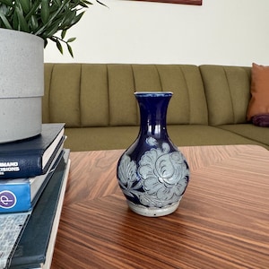 May include: A dark blue ceramic vase with a floral design in light gray. The vase has a bulbous body and a narrow neck. The vase is sitting on a wooden table.