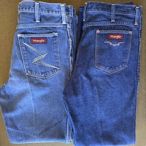 May include: Two pairs of blue denim Wrangler jeans. One pair features a zig-zag design on the back pocket, while the other has a simple pocket design. Both pairs have the Wrangler logo on the back pocket.