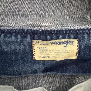 May include: Close-up of a denim jacket's interior, showcasing a rectangular tag. The tag features the Wrangler logo, size details, and care instructions. The jacket's fabric is a medium-wash blue denim.
