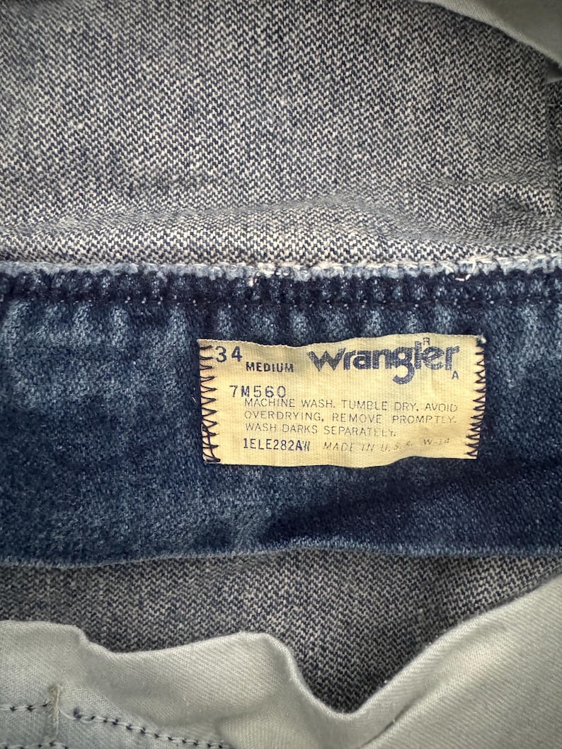 May include: Close-up of a denim jacket's interior, showcasing a rectangular tag. The tag features the Wrangler logo, size details, and care instructions. The jacket's fabric is a medium-wash blue denim.