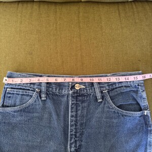 May include: Close-up of blue denim jeans with a measuring tape across the waist. The waist measures approximately 16 inches across. The jeans have classic five-pocket styling and a button and zipper closure.