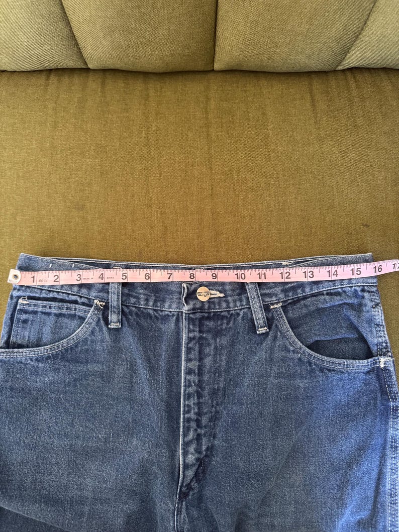 May include: Close-up of blue denim jeans with a measuring tape across the waist. The waist measures approximately 16 inches across. The jeans have classic five-pocket styling and a button and zipper closure.