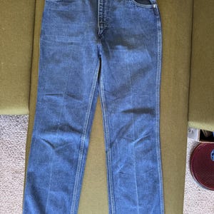 May include: Blue denim jeans with a classic five-pocket design. The jeans have a button and zipper closure, belt loops, and a straight leg cut. The denim is a medium wash, with visible stitching details. The jeans are displayed on a green surface.