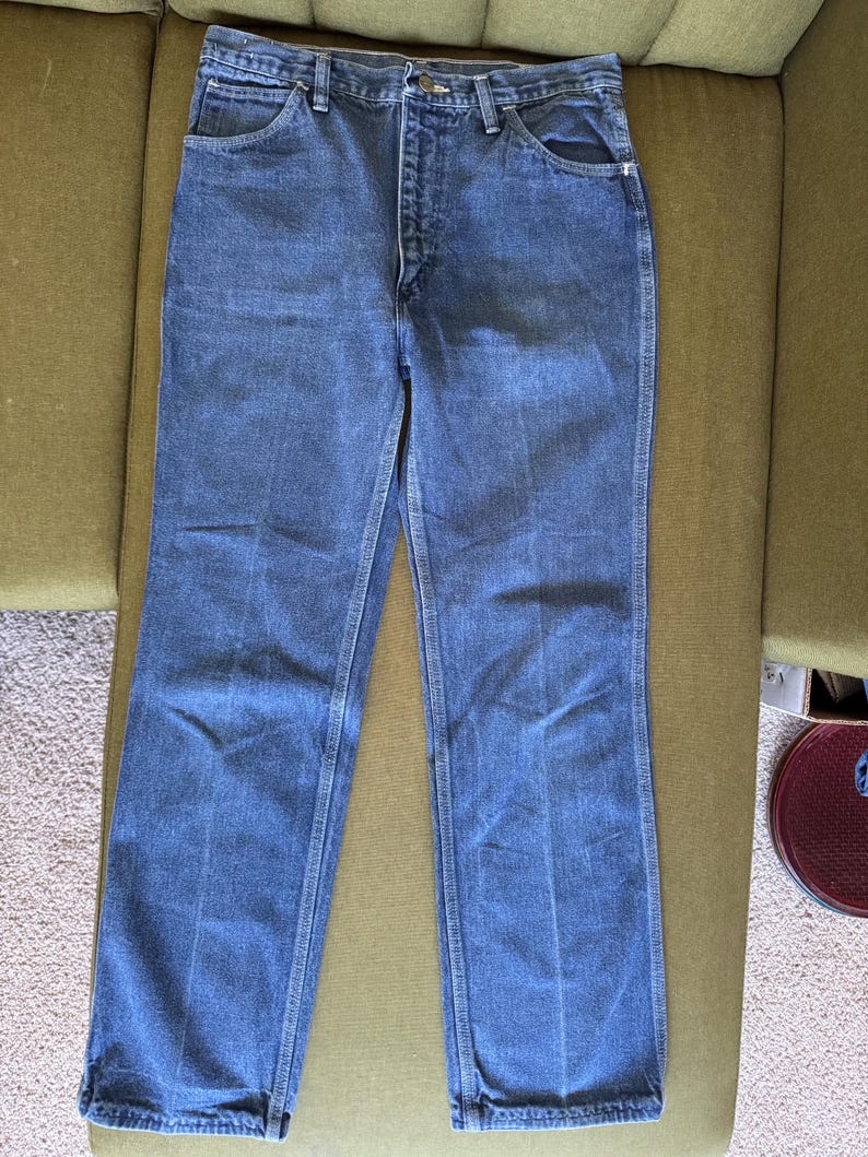 May include: Blue denim jeans with a classic five-pocket design. The jeans have a button and zipper closure, belt loops, and a straight leg cut. The denim is a medium wash, with visible stitching details. The jeans are displayed on a green surface.