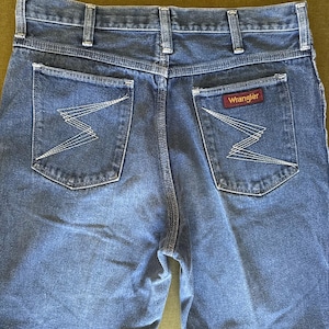 May include: Close-up of the back of blue denim jeans. The jeans have two back pockets with a white zigzag stitch design. A red and white Wrangler label is visible on the right pocket. The jeans have a classic, straight-leg fit.