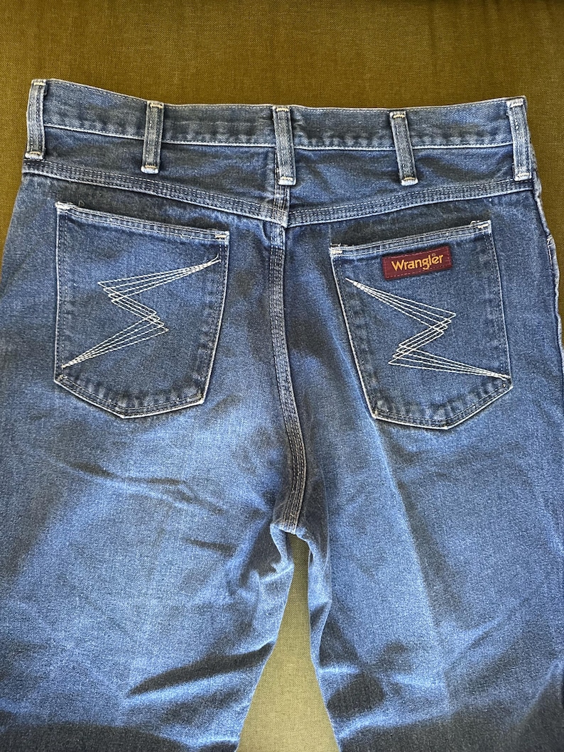 May include: Close-up of the back of blue denim jeans. The jeans have two back pockets with a white zigzag stitch design. A red and white Wrangler label is visible on the right pocket. The jeans have a classic, straight-leg fit.