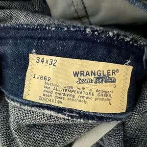 May include: Close-up of blue denim Wrangler jeans. The tag reads "34x32" and "Jeans For Men". The tag also includes washing instructions. The jeans are made of a durable denim fabric.