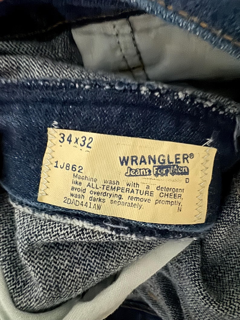May include: Close-up of blue denim Wrangler jeans. The tag reads "34x32" and "Jeans For Men". The tag also includes washing instructions. The jeans are made of a durable denim fabric.