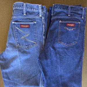 May include: Two pairs of Wrangler denim jeans. One pair is a lighter wash with a zig-zag design on the back pocket, and the other is a darker wash with a stitched design. Both have the Wrangler logo on the back pocket.