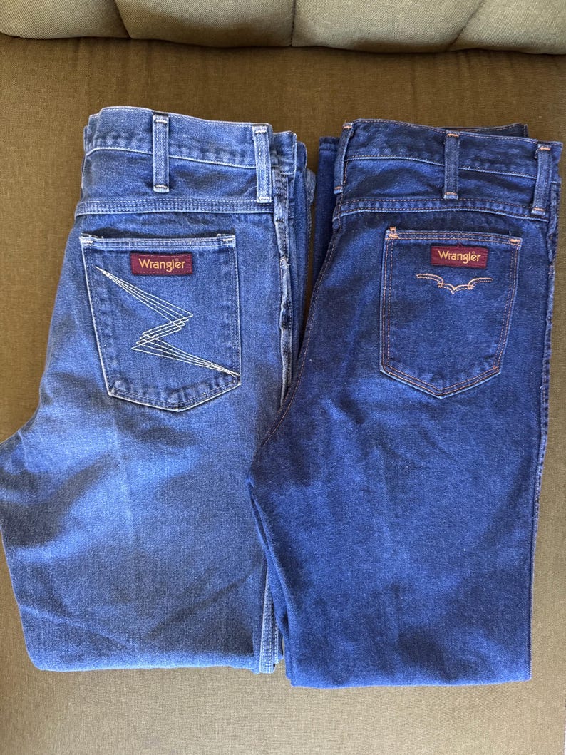 May include: Two pairs of Wrangler denim jeans. One pair is a lighter wash with a zig-zag design on the back pocket, and the other is a darker wash with a stitched design. Both have the Wrangler logo on the back pocket.