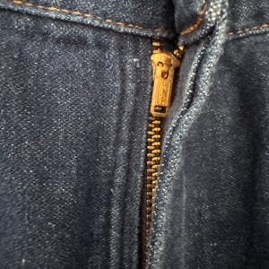 May include: Close-up of a blue denim fabric with a brass zipper. The zipper is partially open, revealing the teeth and pull tab. The denim has a textured appearance with visible stitching.
