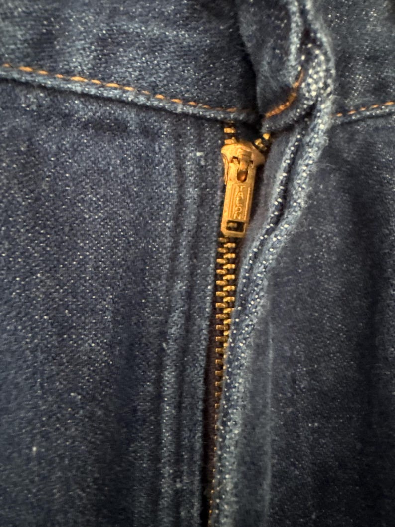May include: Close-up of a blue denim fabric with a brass zipper. The zipper is partially open, revealing the teeth and pull tab. The denim has a textured appearance with visible stitching.