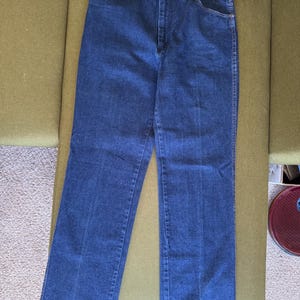 May include: Dark blue denim jeans with a classic five-pocket design. The jeans are straight-legged and have a button and zipper closure. The jeans are laid flat on a green surface.