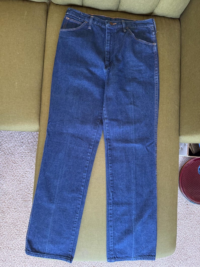 May include: Dark blue denim jeans with a classic five-pocket design. The jeans are straight-legged and have a button and zipper closure. The jeans are laid flat on a green surface.