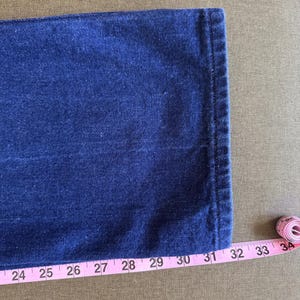 May include: Close-up of a section of dark blue denim jeans. A pink measuring tape is placed along the edge of the jeans, showing measurements from 24 to 34 inches. The jeans have a classic, clean design.