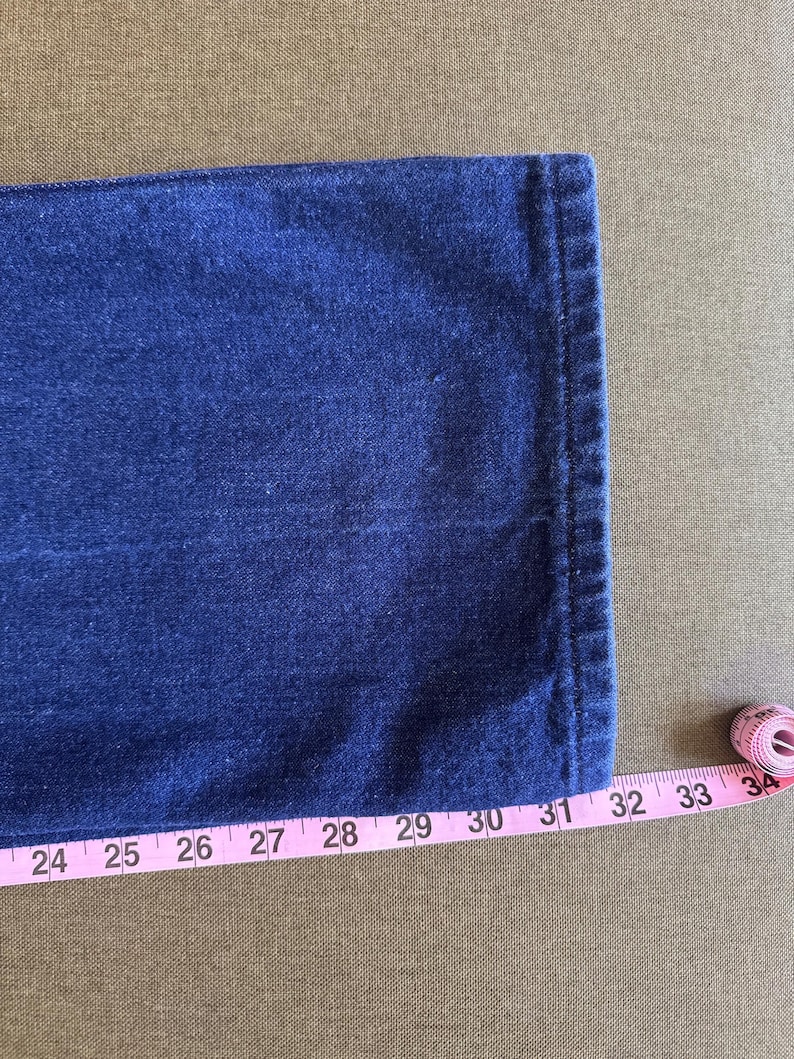 May include: Close-up of a section of dark blue denim jeans. A pink measuring tape is placed along the edge of the jeans, showing measurements from 24 to 34 inches. The jeans have a classic, clean design.