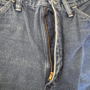 May include: Close-up of blue denim jeans with a partially unzipped brass zipper. The jeans have classic five-pocket styling with belt loops and a button closure. The denim fabric shows subtle fading and wear.