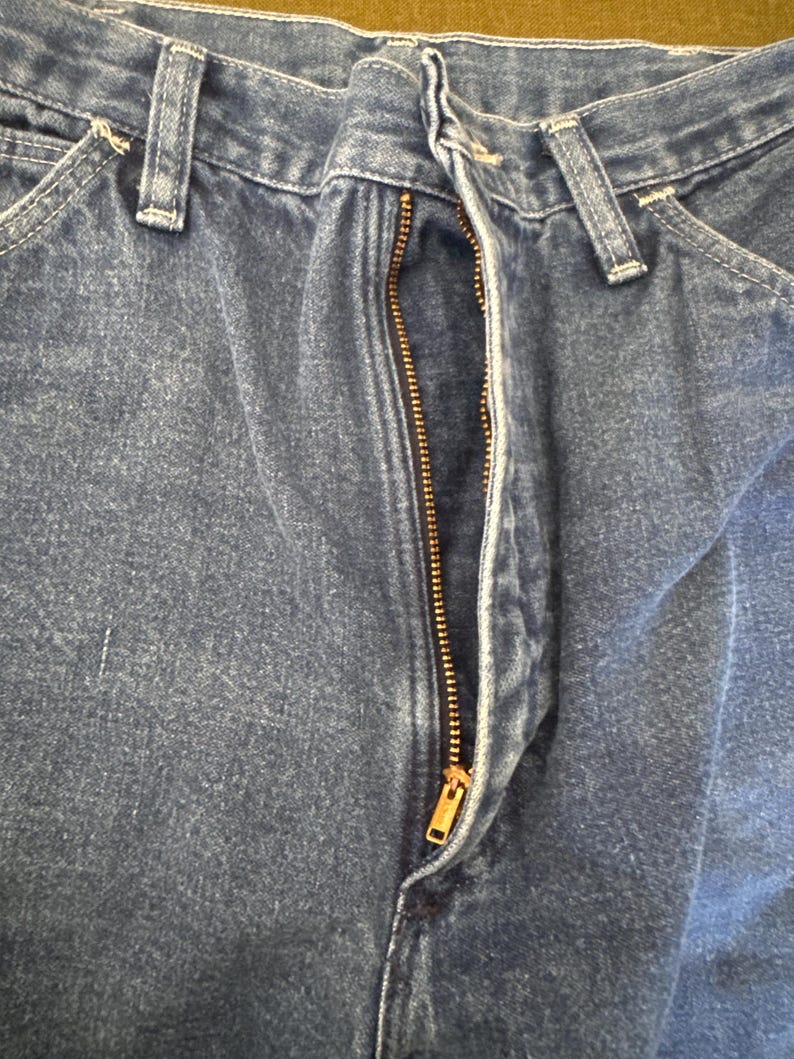 May include: Close-up of blue denim jeans with a partially unzipped brass zipper. The jeans have classic five-pocket styling with belt loops and a button closure. The denim fabric shows subtle fading and wear.