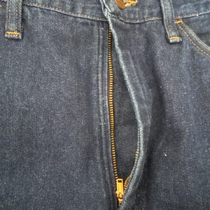 May include: Close-up of dark blue denim jeans with a visible brass zipper and a button closure. The jeans have classic five-pocket styling with reinforced stitching in a contrasting color. The fabric shows subtle wear.