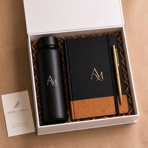 May include: A boxed gift set containing a black water bottle, a black notebook with a cork base, and a gold pen, all bearing a gold "AM" logo. A small card with text is also included in the white box.