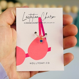 May include: A white card with the text "Lactation Charm" and "to remind yourself which side you are on". The card features a pink bra with a small blue charm hanging from it. The text "HOLLIDAY.CO" is at the bottom of the card.