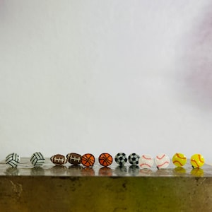 May include: A collection of sports-themed stud earrings. The earrings include designs of a volleyball, football, basketball, soccer ball, baseball, and softball. Each earring is a miniature version of the respective sports ball, with detailed color and design.