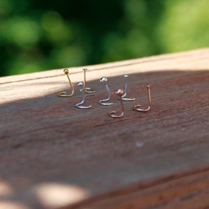 May include: A collection of nose studs in various metallic finishes, including gold, silver, and rose gold. Each stud features a small, decorative top, resting on a wooden surface. The studs are designed with a curved post for secure placement.