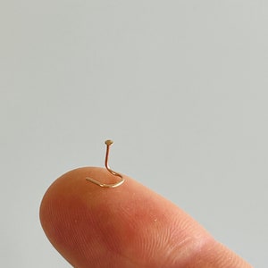 May include: A tiny, gold-colored nose stud with a curved post and a small, round top. The jewelry is shown on a fingertip against a plain, light background. The stud is a delicate piece of body jewelry.