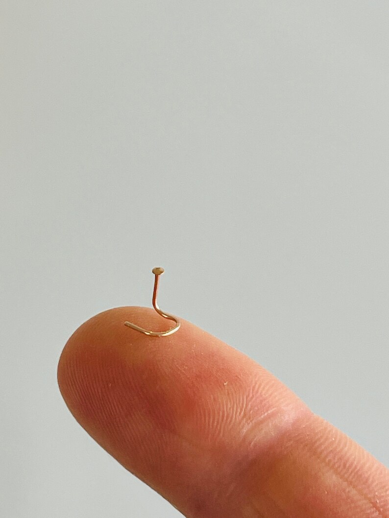 May include: A tiny, gold-colored nose stud with a curved post and a small, round top. The jewelry is shown on a fingertip against a plain, light background. The stud is a delicate piece of body jewelry.