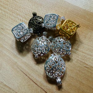 May include: A collection of seven metal cage pendants in silver, gold, and dark bronze tones. These hollow, spherical and cube-shaped charms feature intricate, swirling filigree cutouts throughout their surfaces, designed to hold small items.
