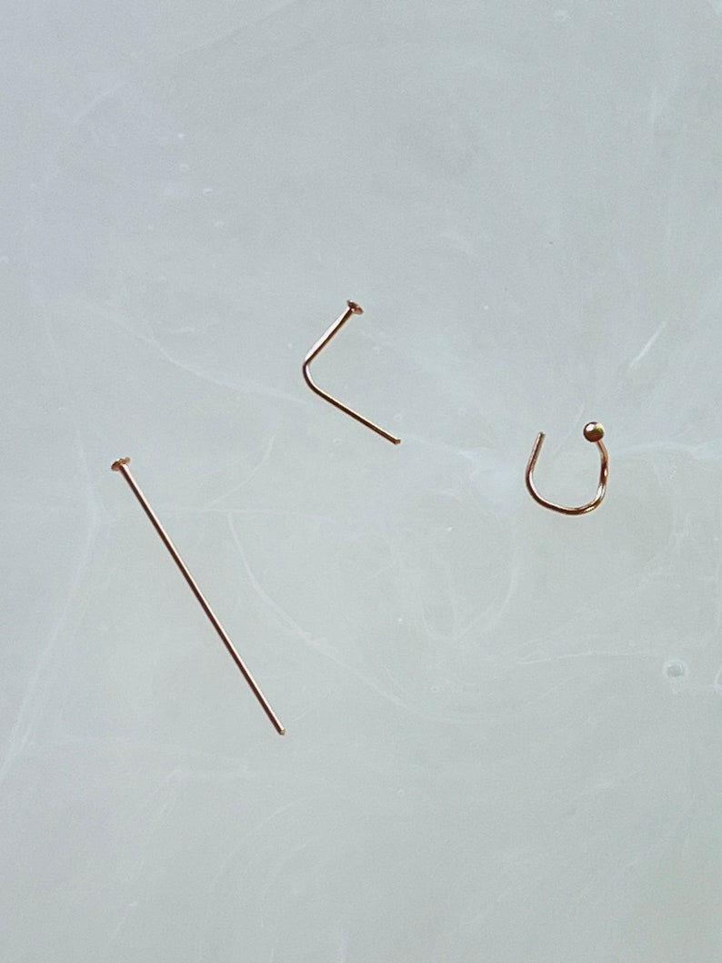 May include: Three gold-colored nose jewelry pieces are displayed against a light background. The pieces include a straight nose stud, a L-shaped nose stud, and a nose ring with a small ball.